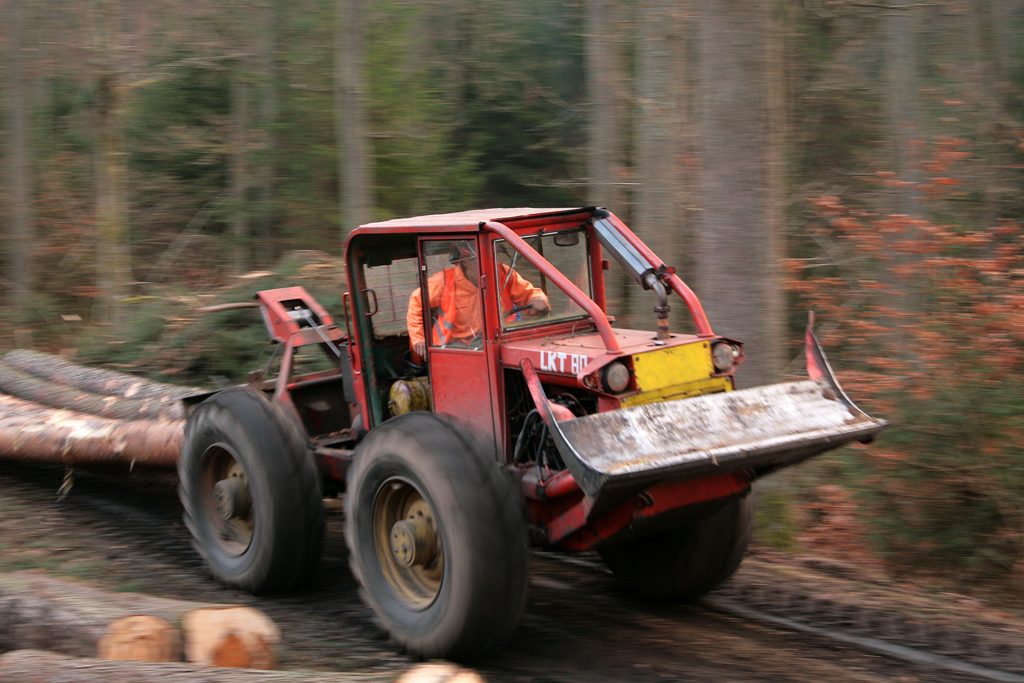 Na zdjęciu widać czerwony skider, maszynę zrywkową, ciągnący półpodwieszone dłużyce. W maszynie znajduje się operator w pomarańczowym stroju roboczym.