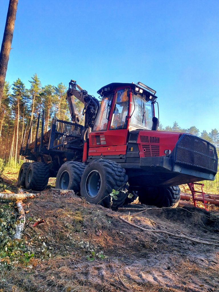 Na zdjęciu widać czerwony forwarder, maszynę zrywkową. Zdjęcie robione "od dołu" co pogłębia wrażenie wielkości i majestatyczności maszyny.