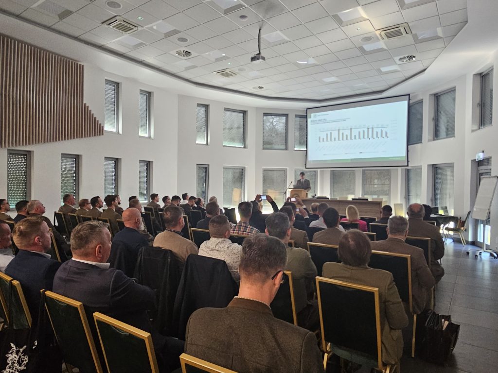 Na zdjęciu widać salę konferencyjną wypełnioną uczestnikami seminarium. Przy drewnianej mównicy stoi mężczyzna w mundurze. Tuż obok mównicy ustawiony jest niewielki stół. Na ekranie za stołem i mównicą wyświetlany jest slajd prezentacji prelegenta na którym widnieją wykresy.