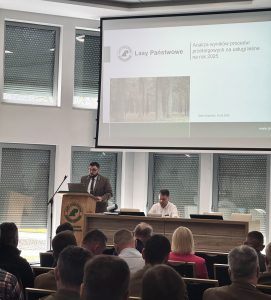 Na zdjęciu widać salę konferencyjną wypełnioną uczestnikami seminarium. Przy drewnianej mównicy stoi mężczyzna w mundurze. Tuż obok mównicy ustawiony jest niewielki stół, przy którym siedzi mężczyzna w białej koszuli. Na ekranie za stołem i mównicą wyświetlany jest slajd prezentacji.