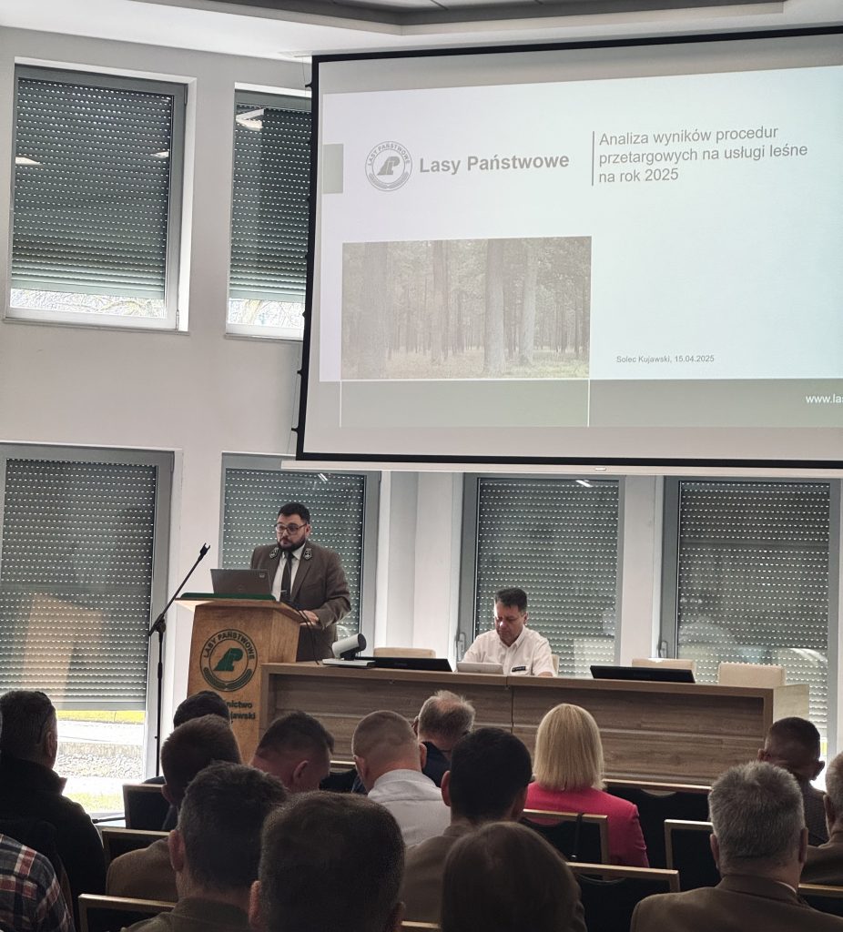 Na zdjęciu widać salę konferencyjną wypełnioną uczestnikami seminarium. Przy drewnianej mównicy stoi mężczyzna w mundurze. Tuż obok mównicy ustawiony jest niewielki stół, przy którym siedzi mężczyzna w białej koszuli. Na ekranie za stołem i mównicą wyświetlany jest slajd prezentacji.