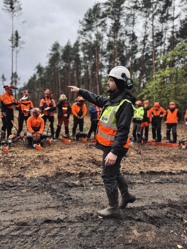 Na obrazku na pierwszym planie widać sędziego zawodów, który zwraca się do zawodników stojących przed nim. Sceneria leśna.