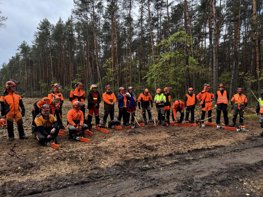 Na zdjęciu widzimy wszystkich zawodników zawodów drwali podczas pierwszego dnia zawodów. Są oni przed rozpoczęciem rywalizacji. Ubrani w odzież ochronną, mają ze sobą pilarki.