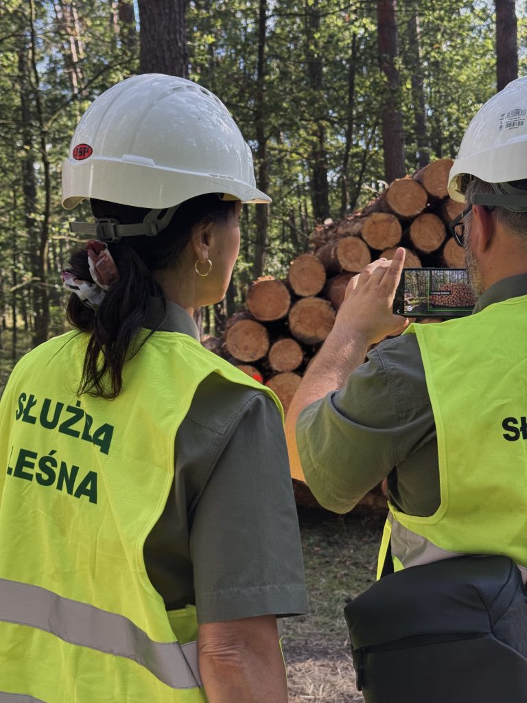 Na zdjęciu widać dwóch pracowników Służby Leśnej. Znajdują się oni w lesie, przed stosem drewna i wykonują pomiar miąższości drewna metodą fotooptyczną.