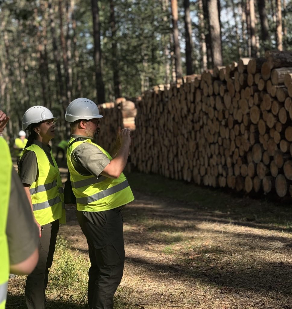 Na zdjęciu widać dwóch pracowników służby leśnej, ubranych w kask i kamizelki odblaskowe. Wykonują pomiar stosu metodą fotooptyczną.